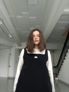 Jenni Österlund with medium length brown hair, white shirt and black dress in front of a stair case