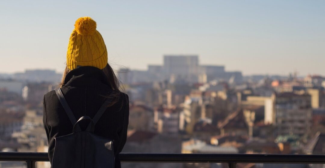 A person wearing a yellow knit hat and black coat stands with their back to the camera, looking out over a cityscape from an elevated viewpoint on a clear day.