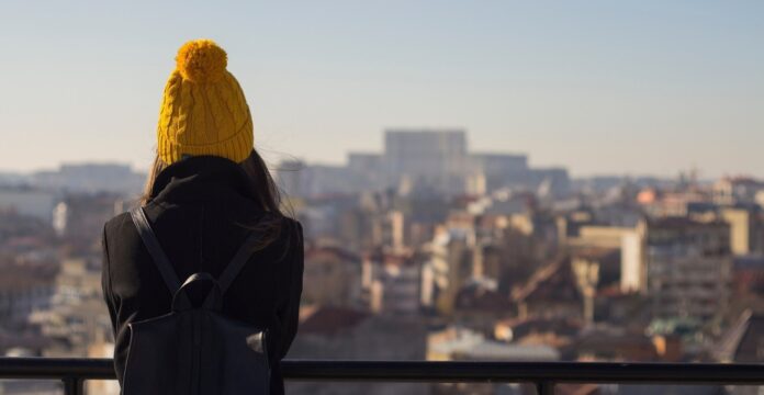 A person wearing a yellow knit hat and black coat stands with their back to the camera, looking out over a cityscape from an elevated viewpoint on a clear day.