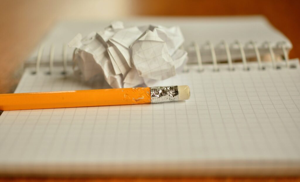  A yellow pencil with a worn eraser lies on a grid-lined spiral notebook, with a crumpled piece of paper resting on the notebook in the background. 